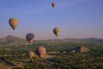 Multi-coloured hot air balloons fly in the sky over a green landscape.