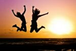 Two people are pictured as sillouhettes jumping into the air by the sea at sunset