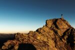 [A person stands at the top of a brown rocky mountain at sunset]