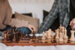 Two people sitting on the sofa playing chess. The chess board is in focus at the forefront of the image, and the two players are out of focus behind.