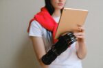 A person holding a tablet with both hands, one of which is a sleek black prosthetic hand. They are wearing a white shirt with a bright red sweater draped over their shoulders.